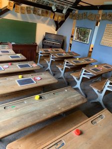 Ein Klassenzimmer aus den 50er Jahren aus der Sicht eines Schülers. Die Schüler sitzen geordnet nach der Klasse auf Holzbänken vor alten Holztischen. In der rechten Ecke des Raumes kann man ein Klavier sehen, auf dem die Lehrer ab und zu Musik spielen.