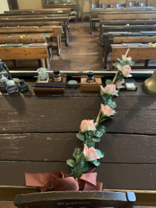 Mit Blumen verzierter Rohrstock auf einem Lehrerpult. Im Hintergrund sind die Holzbänke der Schüler zu sehen. Die Schüler tuen dies üblicherweise am Geburtstag des Lehrers