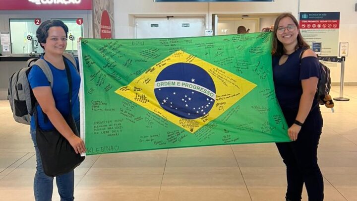 Zwei junge Frauen stehen in einer Flughafen-Abflughalle und halten eine brasilianische Flagge.