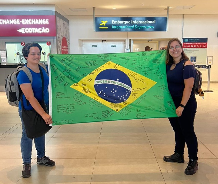 Zwei junge Frauen stehen in einer Flughafen-Abflughalle und halten eine brasilianische Flagge.