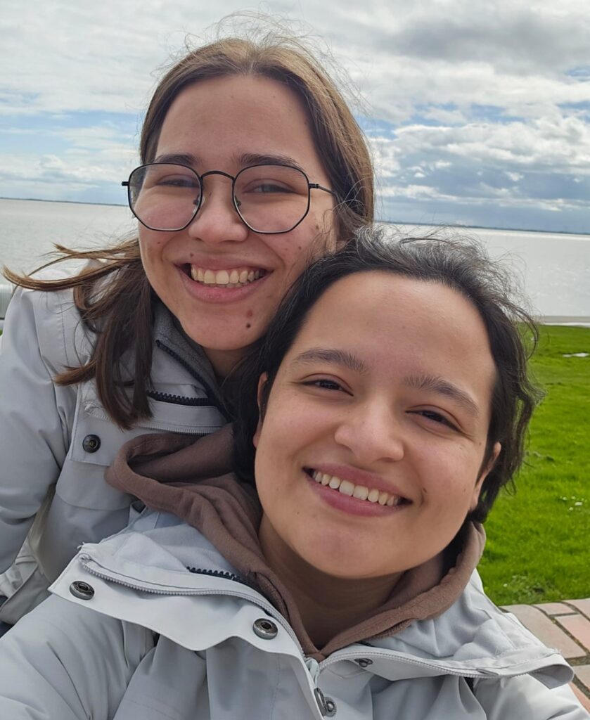 Zwei junge Frauen lächeln in die Kamera. Im Hintergrund Wasser und Wiese am Wilhelmshavener Südstrand.