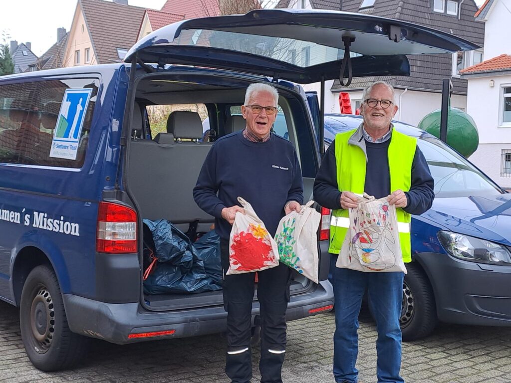 Günter Kallweit und Jürgen Benecken stehen beim Kofferraum eines Autos und halten Taschen in die Luft.