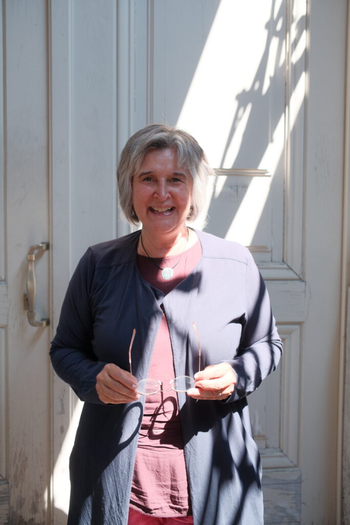 Maria Noichl steht vor einer weißen Tür und lächelt in die Kamera. Sie hält ihre Brille in der Hand.